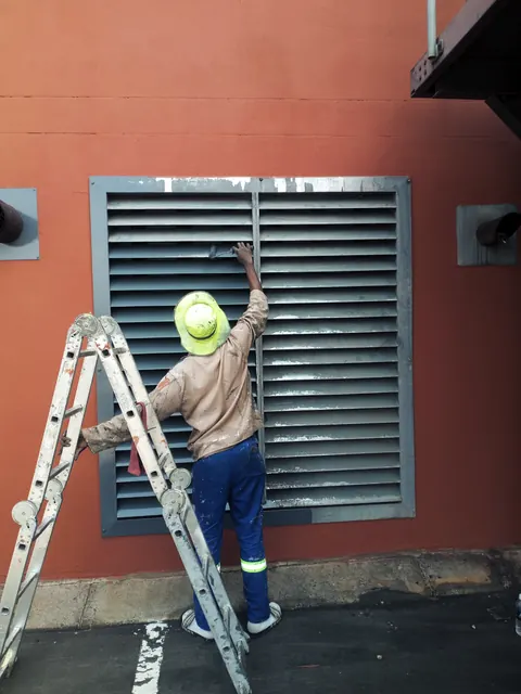 Painter on a ladder painting a steel louvre vent on an orange wall