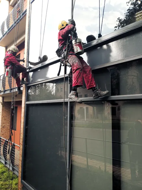 Two MP technicians on ropes repainting a dark steel fascia on an apartment block