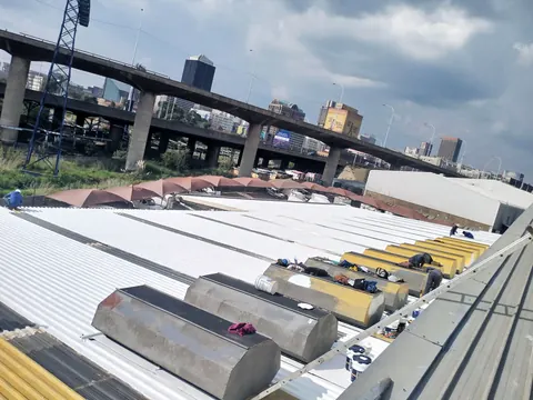 MP crew coating a factory roof white, with the Johannesburg skyline and a highway bridge behind