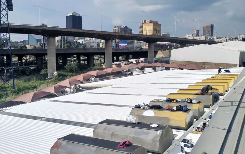 MP crew coating a factory roof white, with the Johannesburg skyline and a highway bridge behind